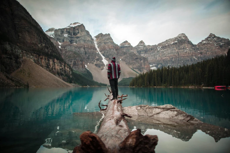 a man standing on a log in front of a lake, inspired by Michael Komarck, pexels contest winner, banff national park, monica langlois, conde nast traveler photo, mountain range