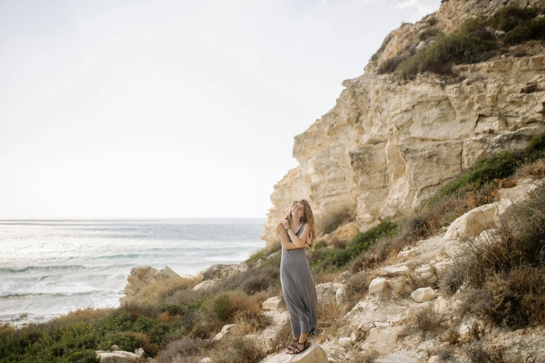 a woman standing on top of a cliff next to the ocean, by Simon Marmion, happening, long dress, cyprus, portrait image