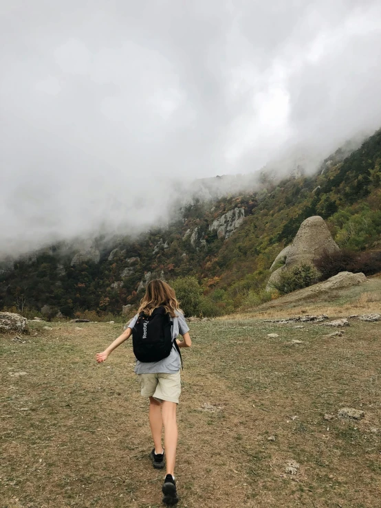 a woman with a backpack walking up a hill, a picture, by Lucia Peka, cloudy weather, rocky foreground, profile image