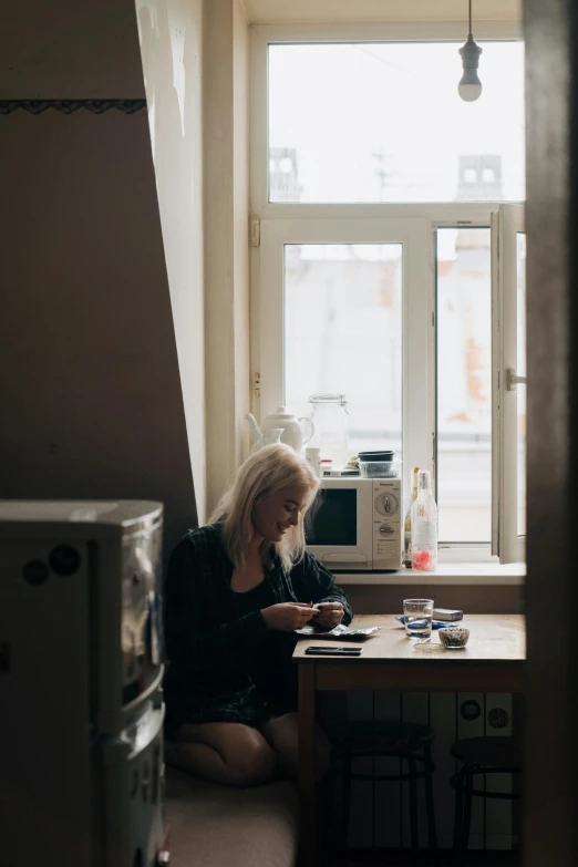 a woman sitting at a table in front of a window, a polaroid photo, inspired by Elsa Bleda, unsplash contest winner, small kitchen, russian girlfriend, a blond, inside an old apartment