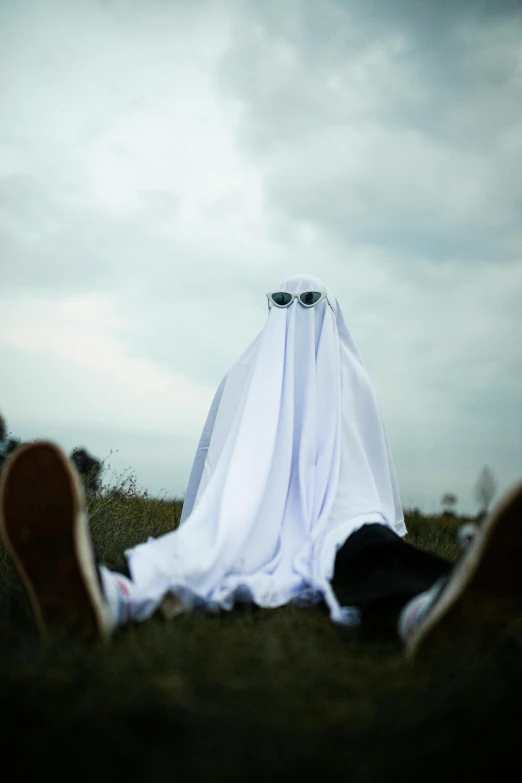 a person sitting on top of a grass covered field, halloween ghost under a sheet, wearing white suit and glasses, ( ( theatrical ) ), shroud