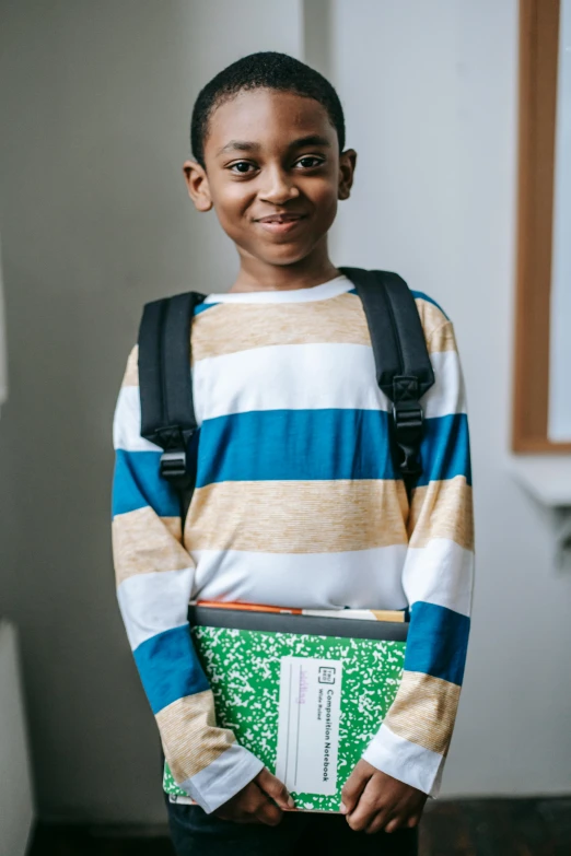 a young boy standing in a hallway holding a book, square backpack, wearing a green sweater, jemal shabazz, wearing stripe shirt