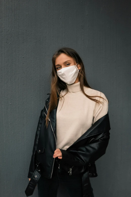 a woman wearing a face mask standing against a wall, a picture, trending on pexels, wearing a turtleneck and jacket, synthetic, beige, dark backdrop