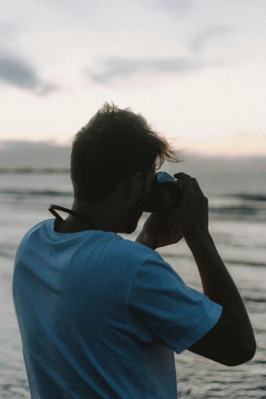 a man standing on top of a beach next to the ocean, inspired by Max Dupain, unsplash contest winner, holding a dslr camera, profile shot, dusk setting, close-up shot from behind