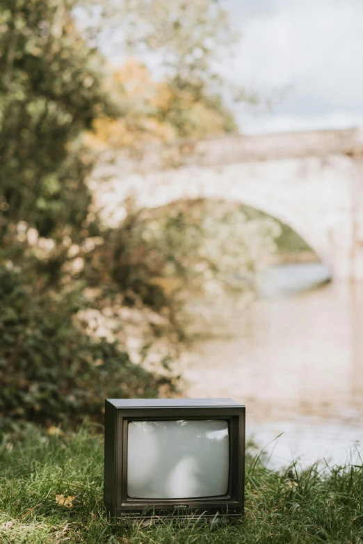 an old television sitting in the grass next to a river, inspired by Camille Corot, unsplash, video art, sitting under bridge, autumnal, vertical movie frame, grey