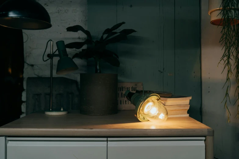 a lamp sitting on top of a wooden table, a still life, inspired by Elsa Bleda, greenish lighting, lightbulb, with one vintage book on a table, tactile buttons and lights