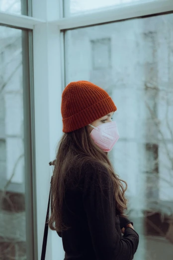 a woman standing in front of a window wearing a face mask, a picture, trending on pexels, wearing a beanie, red hues, profile image, downward somber expression