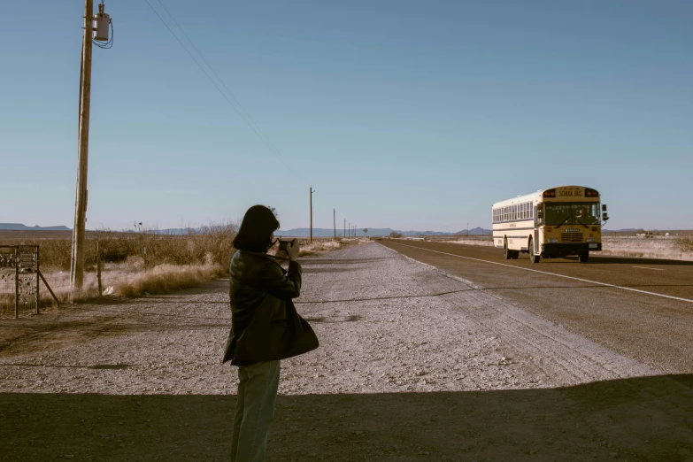 a woman standing on the side of a road next to a bus, a picture, by Kristin Nelson, unsplash, standing in a barren field, movie filmstill, clear skies in the distance, taking a picture