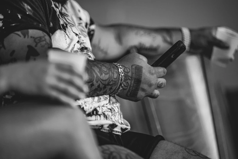 a close up of a person holding a cell phone, a tattoo, by Adam Marczyński, sitting in a lounge, back and white, arms covered in gang tattoo, warm weather