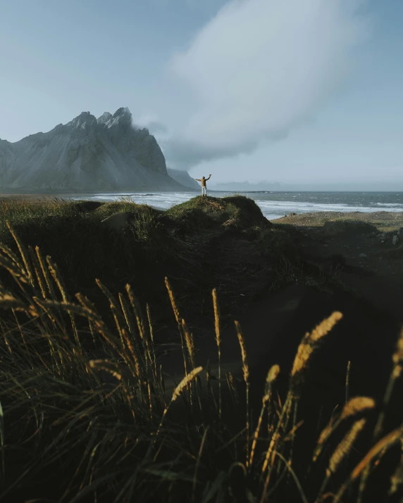 a man standing on top of a cliff next to the ocean, by Johannes Voss, unsplash contest winner, mountain behind meadow, dancing on the beach, standing in tall grass, black sand