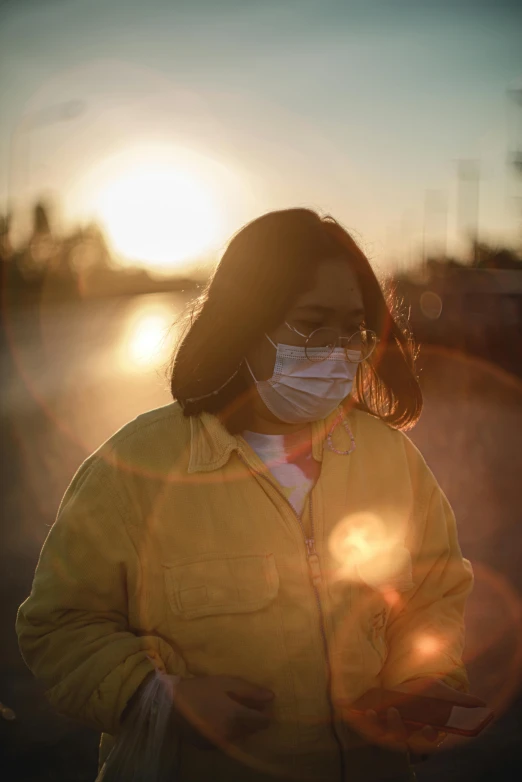 a woman wearing a face mask at sunset, a picture, trending on pexels, wearing a yellow hoodie, leslie zhang, healthcare worker, heavy looking