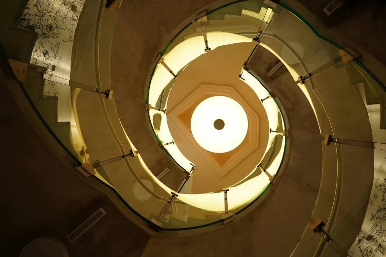 a spiral staircase in a building with glass railings, an album cover, inspired by M.C. Escher, art nouveau, yellow artificial lighting, john pawson, looking down from above, looking up at camera
