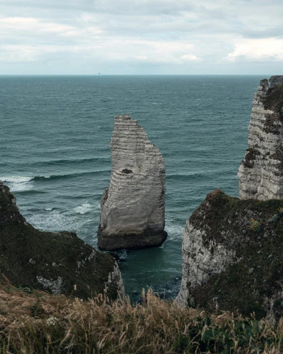 a man standing on top of a cliff next to the ocean, an album cover, by Raphaël Collin, pexels contest winner, structural geology, northern france, he got a big french musctache, subreddit / r / whale