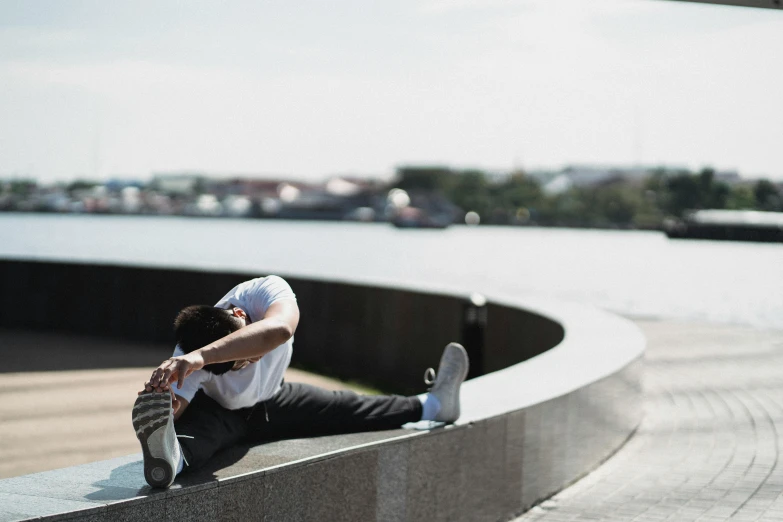 a man sitting on top of a wall next to a body of water, happening, dynamic stretching, sweat and labour, sydney park, promo