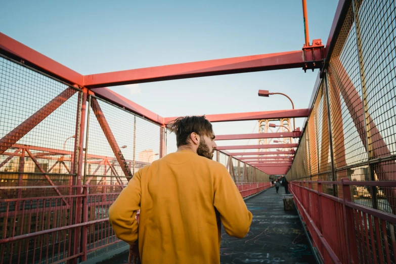 a man in a yellow coat walking across a bridge, an album cover, inspired by Elsa Bleda, pexels contest winner, happening, red trusses, looking over city, long orange sweatshirt, zachary corzine