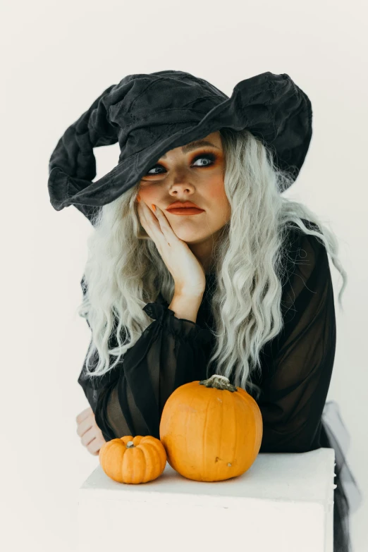 a woman wearing a witch hat and holding two pumpkins, trending on pexels, curled silver hair, sydney sweeney, hand on her chin, ( 3 1