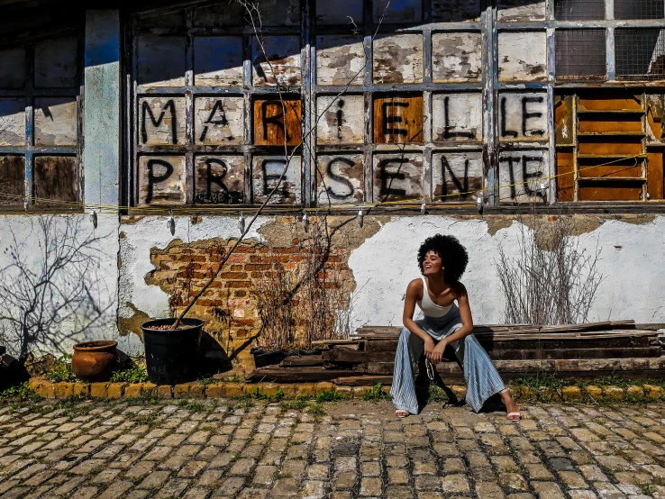 a woman sitting on a bench in front of a building, an album cover, by Andrée Ruellan, pexels contest winner, graffiti, maria borges, while marble, in front of a garage, jacqueline e