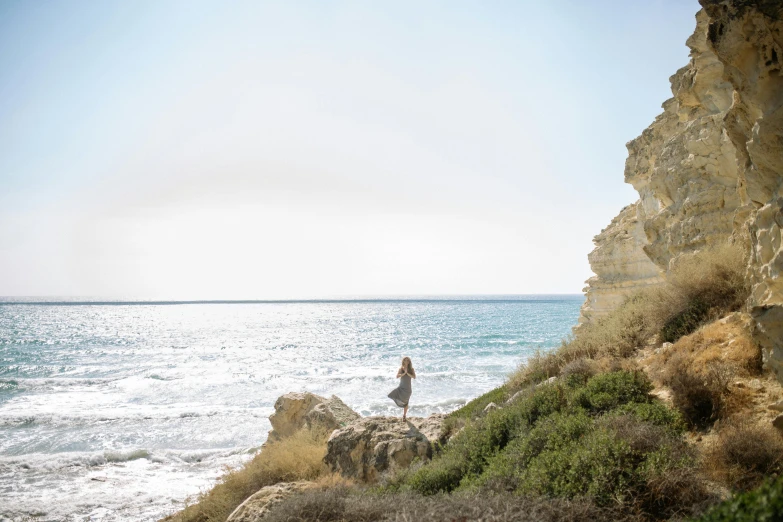 a woman standing on top of a cliff next to the ocean, by Simon Marmion, cyprus, grey, jen atkin, conde nast traveler photo