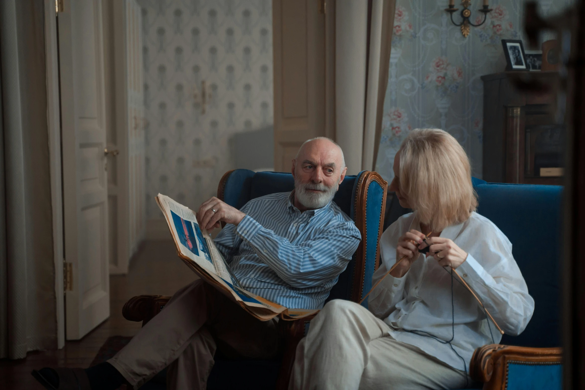 a man and a woman sitting on a blue chair, old gigachad with grey beard, reading glasses, sitting across the room, image