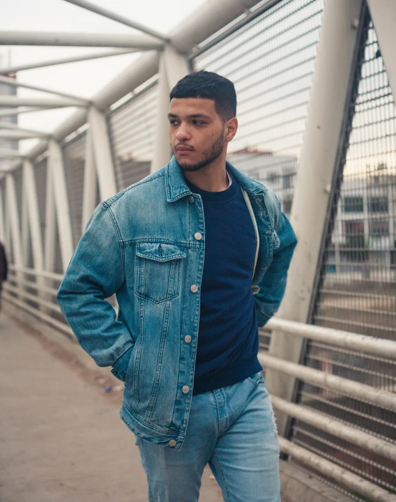 a man standing on a bridge with his hands in his pockets, an album cover, by Cosmo Alexander, trending on pexels, renaissance, wearing double denim, mixed race, model wears a puffer jacket, blue hue