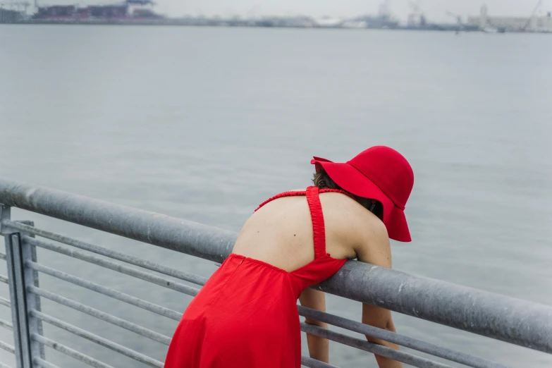 a woman in a red dress leaning on a railing, inspired by Ruth Orkin, pexels contest winner, hat covering eyes, the sea of sadness, red body suit, face down