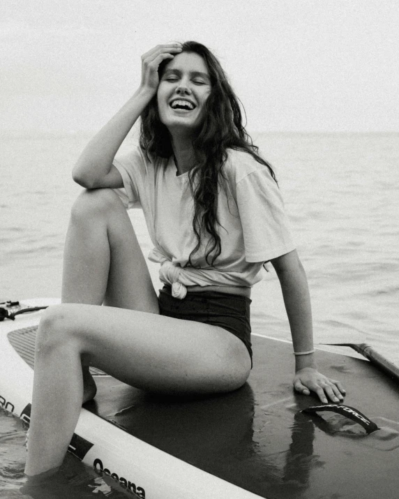 a woman sitting on top of a surfboard in the water, a black and white photo, by Olivia Peguero, trending on unsplash, smiling fashion model, wearing a t-shirt, asher duran, smiling slightly