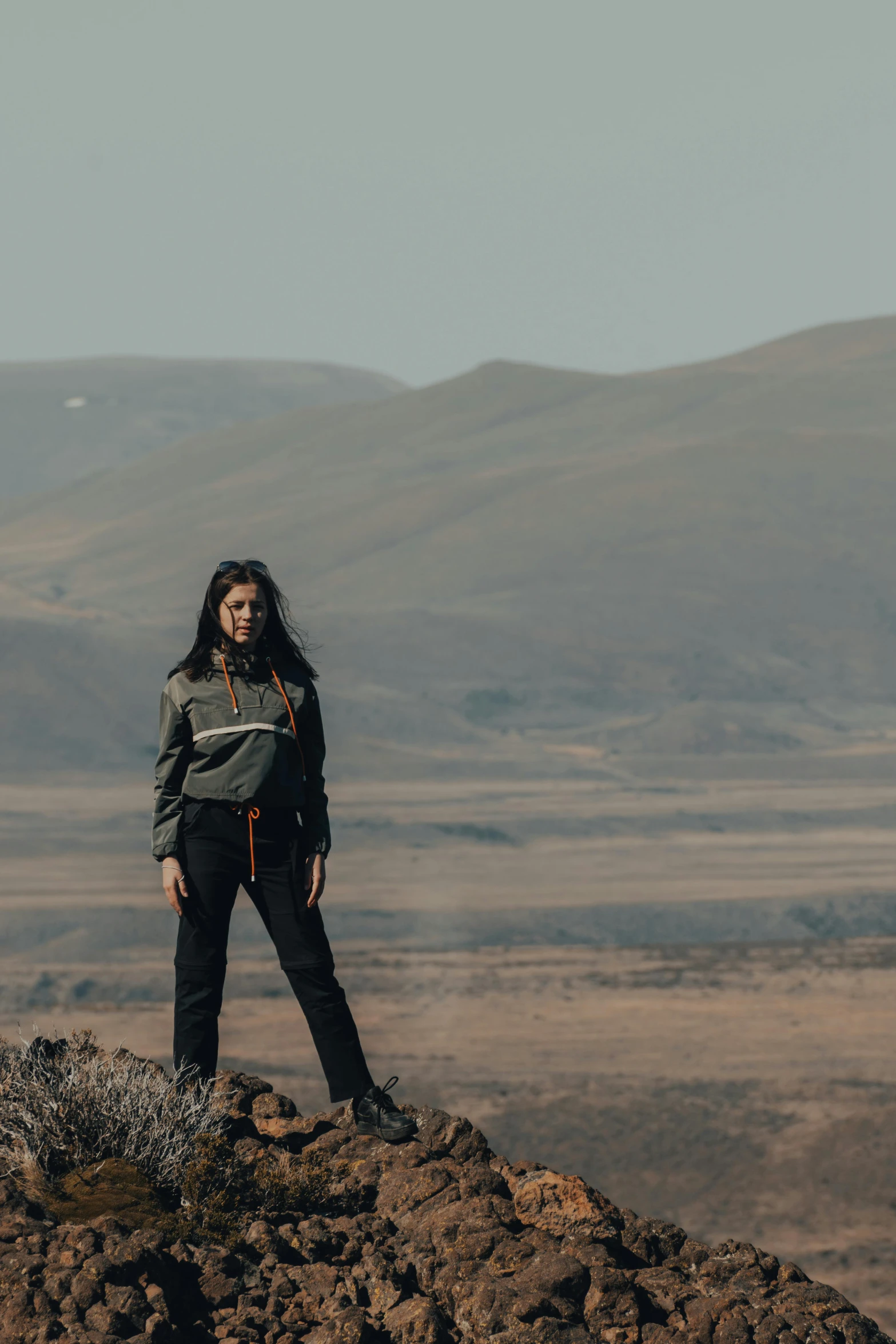 a woman standing on top of a rocky hill, a portrait, by Louisa Matthíasdóttir, trending on unsplash, wearing space techwear, iceland hills in the background, standing in the savannah, background image