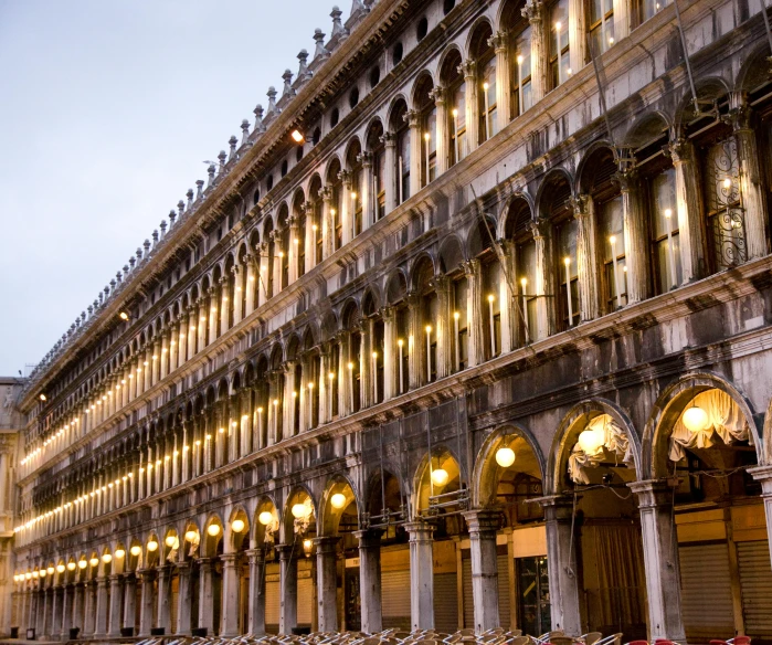 a row of tables and chairs in front of a building, an album cover, inspired by Quirizio di Giovanni da Murano, pexels contest winner, renaissance, venice at dusk, square, many floors, white sweeping arches