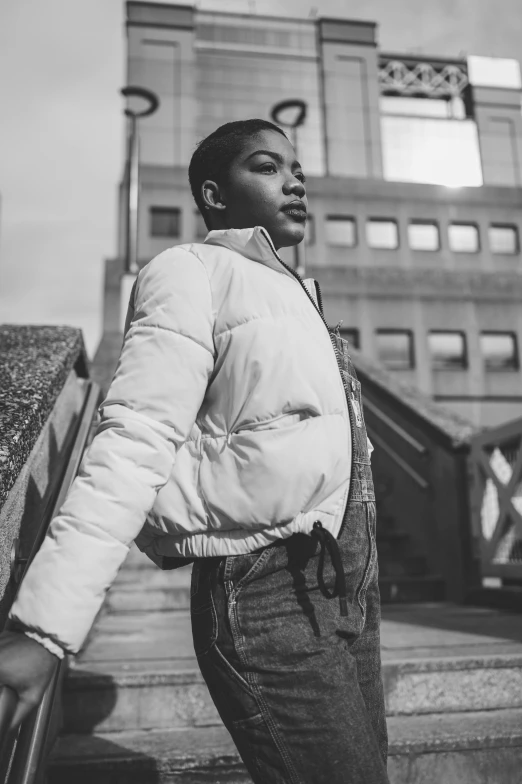 a man standing on the steps of a building, a black and white photo, by Thomas Fogarty, pexels, model wears a puffer jacket, black teenage girl, promotional image, the girl and the sun