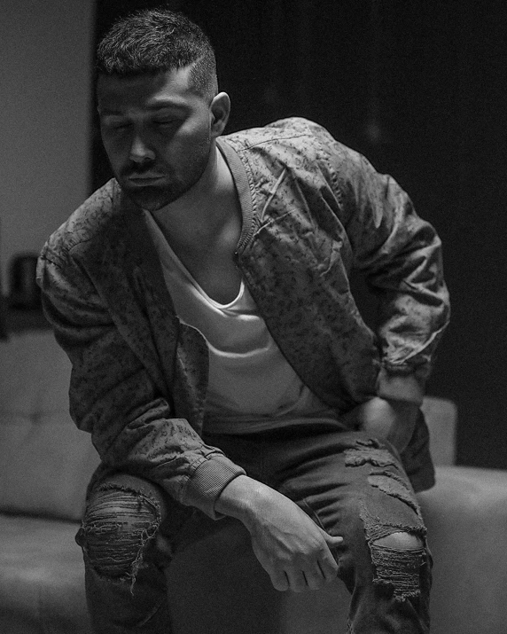 a black and white photo of a man sitting on a couch, by Robbie Trevino, singer maluma, wearing ripped dirty flight suit, sad man, an aviator jacket and jorts