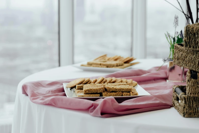 a table topped with plates of food next to a window, inspired by Richmond Barthé, unsplash, private press, marshmallow graham cracker, white and pink cloth, background image, low angle shot