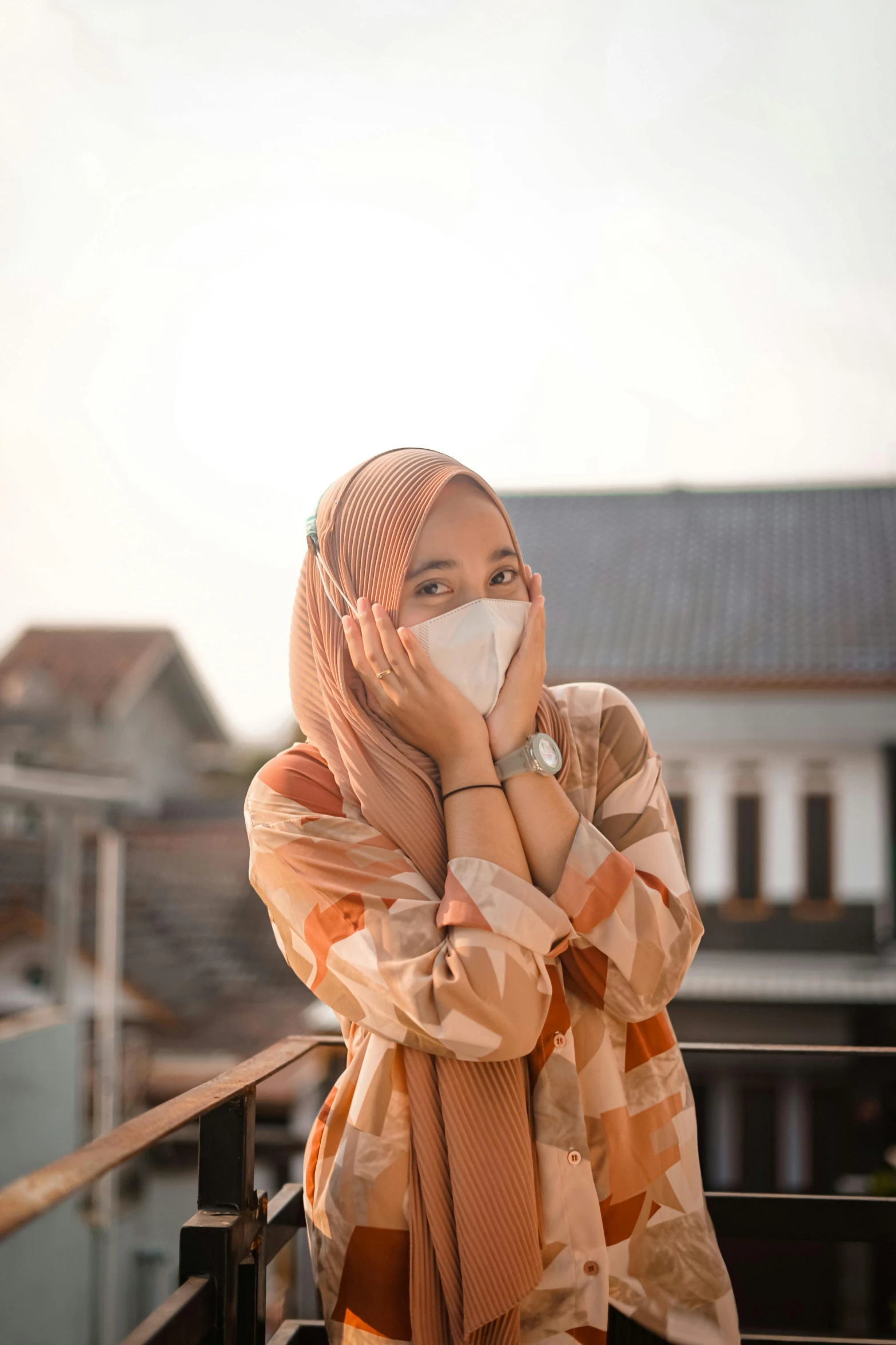 a woman wearing a face mask standing on a balcony, a picture, trending on pexels, hurufiyya, indonesia, hear no evil, blushing, hiding in the rooftops