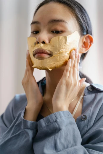 a woman putting a gold facial mask on her face, by Julia Pishtar, cardboard, silicone skin, vietnamese woman, face and body
