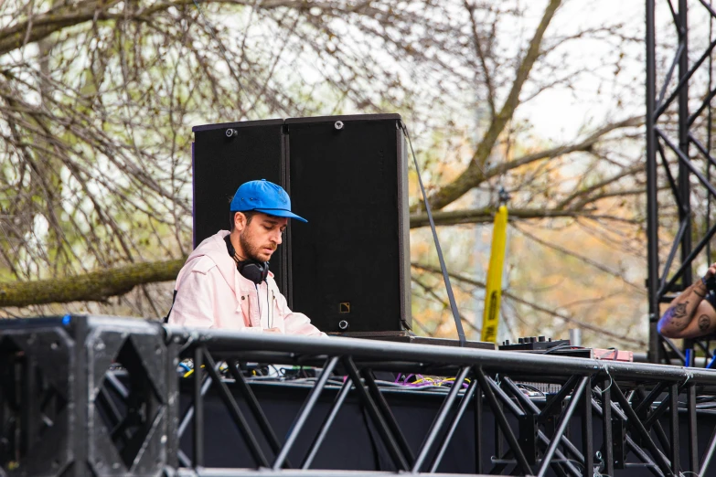 a man sitting at a table with headphones on, by Julia Pishtar, an outdoor festival stage, during spring, against the backdrop of trees, turntables