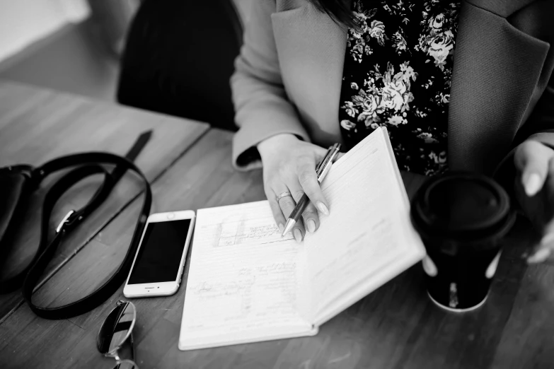 a woman sitting at a table writing on a piece of paper, a black and white photo, pexels, holding notebook, phone photo, thumbnail, fashionable