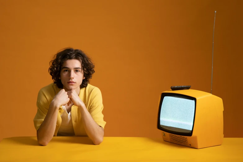 a man sitting at a table in front of a television, an album cover, pexels, yellow clothes, timothee chalamet, retro tv, avatar image