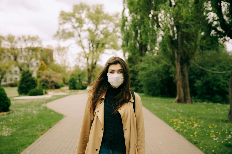 a woman walking down a sidewalk wearing a face mask, a picture, by Emma Andijewska, trending on pexels, renaissance, with a park in the background, gemma chan, polluted, beige