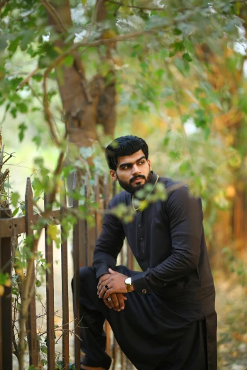 a man sitting on the ground next to a fence, a picture, by Max Dauthendey, pexels contest winner, a portrait of rahul kohli, trees in background, elegant pose, headshot profile picture