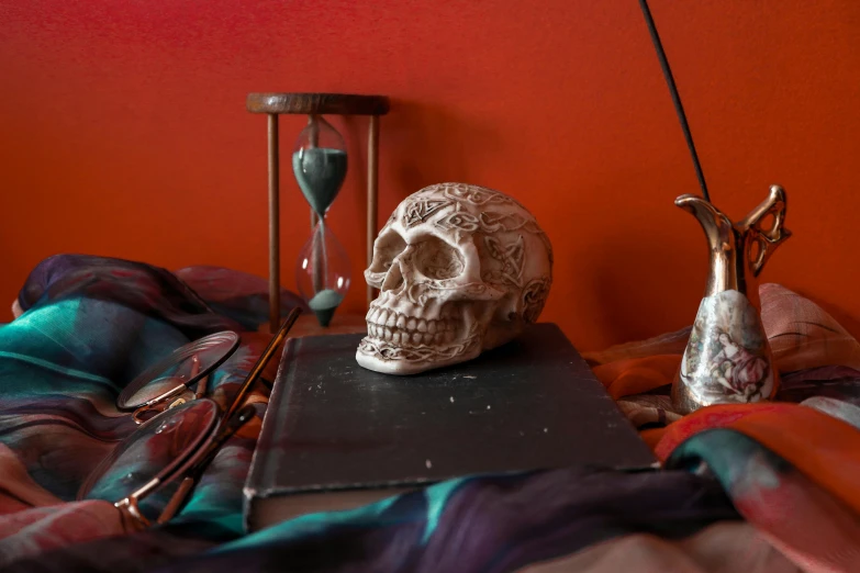 a skull sitting on top of a bed next to a book, a still life, trending on pexels, vanitas, gray and orange colours, tabletop game props, red mood in background, three - quarter view