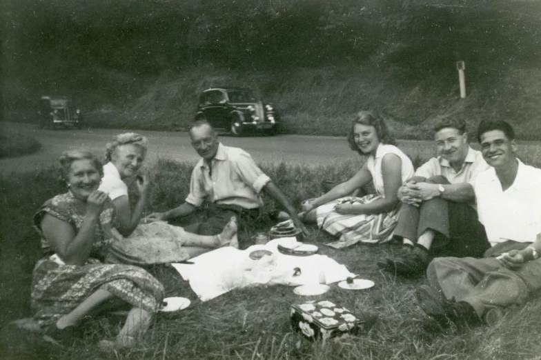 a group of people sitting on top of a grass covered field, a black and white photo, inspired by Bert Hardy, eating outside, on a road, while smiling for a photograph, picnic