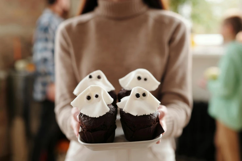 a woman holding a tray of cupcakes with marshmallow ghosts on them, by Helen Stevenson, trending on pexels, white glowing eyes, brown, black, chiffon