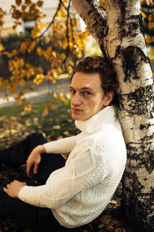 a man sitting on the ground next to a tree, an album cover, inspired by Sergei Sviatchenko, he is wearing a brown sweater, luke evans, pale skin curly blond hair, close - up portrait shot