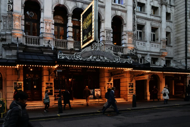 a group of people walking down a street in front of a theater, london architecture, marquee, nomad, brown