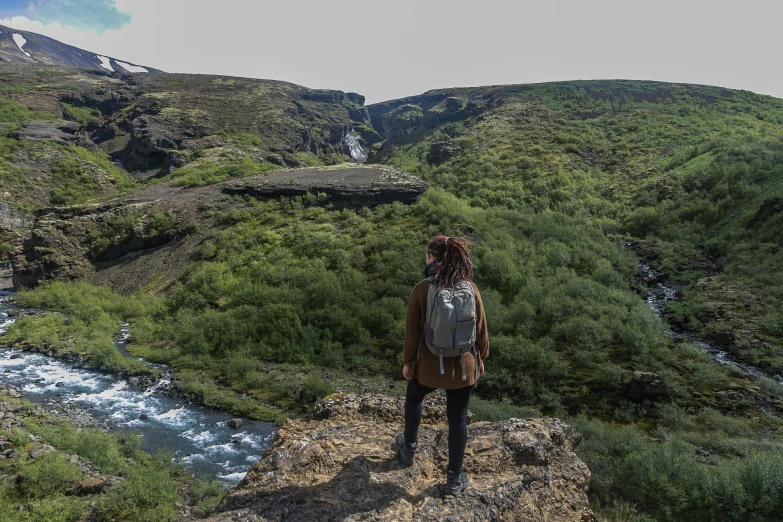 a woman standing on top of a mountain next to a river, by Hallsteinn Sigurðsson, pexels contest winner, hurufiyya, a man wearing a backpack, thumbnail, lush surroundings, facing sideways