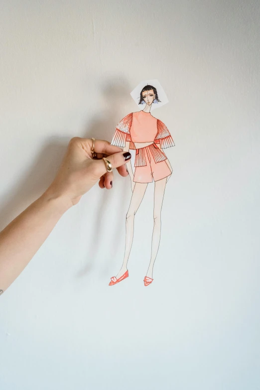 a hand holding a cutout of a woman in a dress, a cartoon, by Olivia Peguero, pexels, technical drawings, wearing an orange jumpsuit, on white paper, on a wall