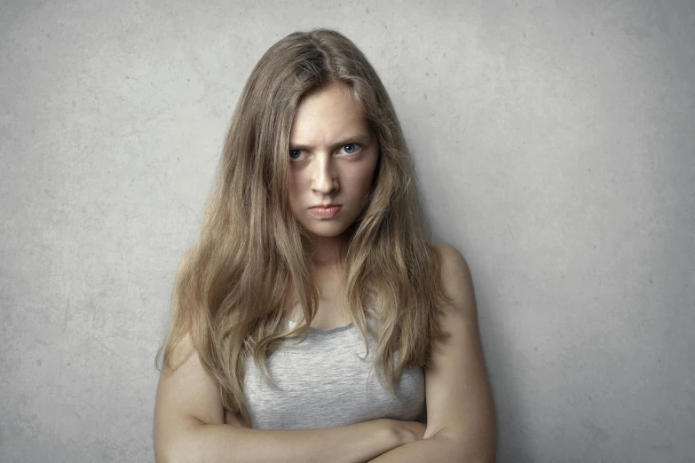 a woman leaning against a wall with her arms crossed, a portrait, trending on pexels, young angry woman, square, grey, fighting