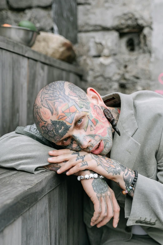 a man with tattoos on his face leaning against a wall, a tattoo, inspired by Seb McKinnon, trending on pexels, hyperrealism, wearing a worn out suit, lying down, hairless, slide show