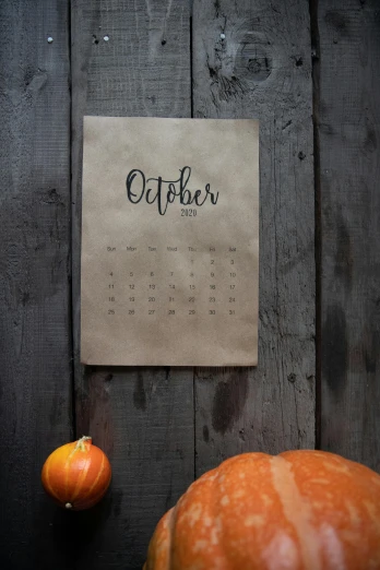 a calendar sitting on top of a wooden table next to a pumpkin, by Julia Pishtar, pexels, square, light - brown wall, made of cardboard, gray
