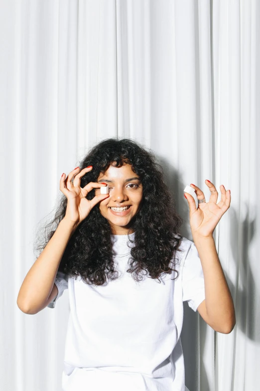 a woman standing in front of a white curtain, by Olivia Peguero, pexels contest winner, curly bangs, offering the viewer a pill, channeling third eye energy, wearing two metallic rings