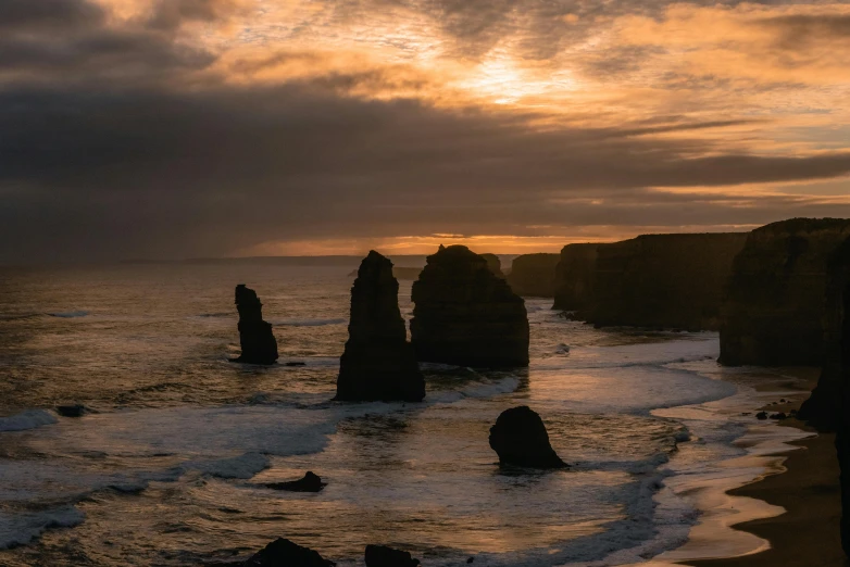 a couple of large rocks sitting on top of a beach, by Andrew Geddes, pexels contest winner, australian tonalism, golden pillars, sunset panorama, majestic spires, roaring ocean in front